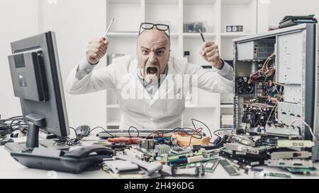 Tecnico arrabbiato frustrato che tenta di riparare un computer, la sua scrivania è piena di parti del computer Foto Stock
