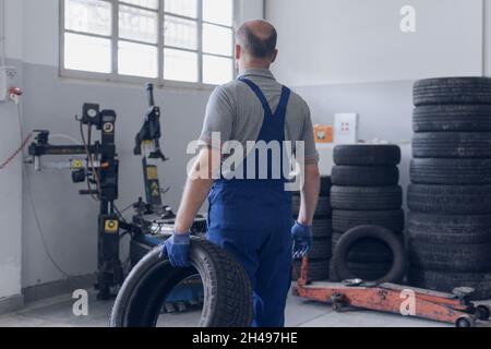 Meccanico professionista che lavora nella stazione di riparazione auto, sta trasportando un pneumatico Foto Stock