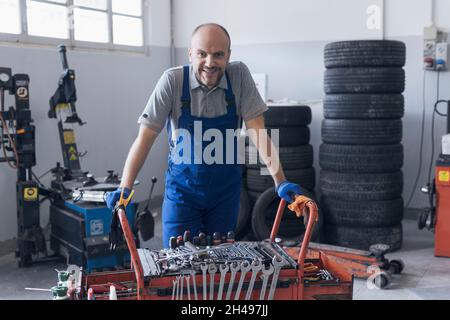 Meccanico che lavora nel negozio di riparazione auto, è appoggiato su un carrello porta attrezzi e sorridente Foto Stock