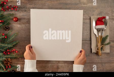 Le mani della donna tiene il menu nelle sue mani mentre si siede al tavolo della cena di Natale. Decorazioni albero di Natale bordo su tavolo in legno d'epoca con posate e Foto Stock