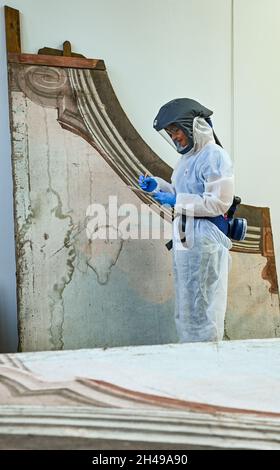 01 novembre 2021, Brandeburgo, Wünsdorf: Un restauratore lavora in un vestito protettivo nel laboratorio di restauro del Landesamt für Denkmalpflege und Archäologisches Landesmuseum (BLDAM) su una pittura a figura di un teatro barocco ambientato dall'Abbazia di Neuzelle. Grazie al generoso sostegno a lungo termine del commissario federale per la cultura e i mezzi di comunicazione (BKM), l'Ostdeutsche Sparkassensstiftung con lo Sparkasse Oder-Spree e la disponibilità del Ministero della scienza, della ricerca e della cultura (MWFK), dell'Ufficio di Stato di Brandeburgo per la conservazione dei monumenti e del Museo Archeologico di Stato Foto Stock