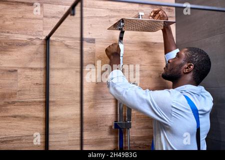 Afro american Professional handyman che lavora in bagno doccia interno, vista laterale ritratto, bel idraulico professionale in tute installazione rubinetto acqua Foto Stock