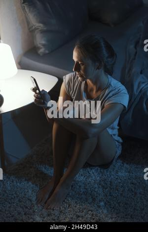 Donna esausta seduta accanto al suo letto e controllando il suo smartphone a tarda notte, sta soffrendo di insonnia Foto Stock