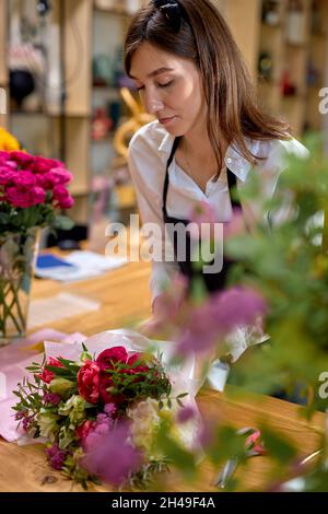 professionista fiorista donna fatto bouquet di fiori di fioritura stagionale, preparandoli per la vendita. Affascinante signora caucasica in grembiule sul posto di lavoro, focalizzata Foto Stock