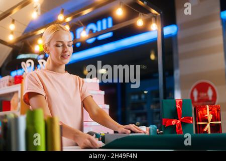 Allegro attraente giovane donna che avvolge la scatola del regalo di Natale di festa in carta artigianale al tavolo del negozio di festa. Foto Stock