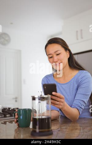 Donna asiatica felice bevendo caffè e utilizzando smartphone in cucina Foto Stock
