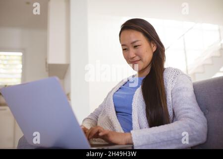Donna asiatica felice seduta sul divano con il computer portatile a casa Foto Stock