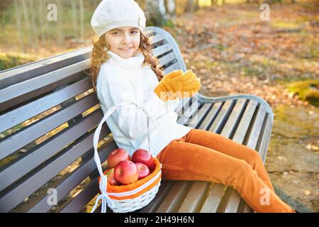 sorridente ragazza in un maglione bianco e berretto si siede su una panchina nel parco in autunno. Foto di alta qualità Foto Stock