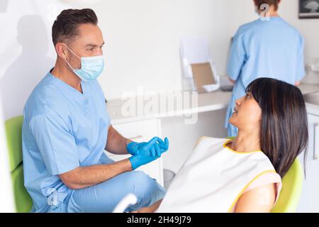 Dentista maschio caucasico che indossa maschera facciale che parla con paziente femminile in clinica dentale moderna Foto Stock