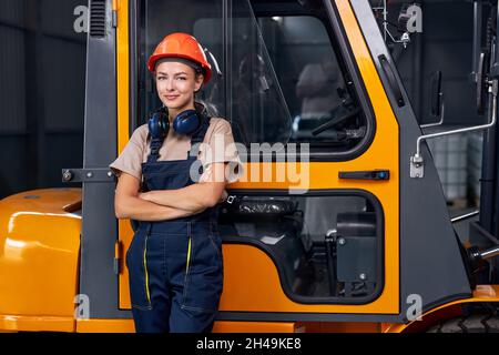 sorridente lavoratore femminile di lavoro vicino a carrello elevatore a forche in fabbrica industria logistica magazzino di spedizione, in posa con le braccia ripiegate, vestito in uniforme e oran Foto Stock