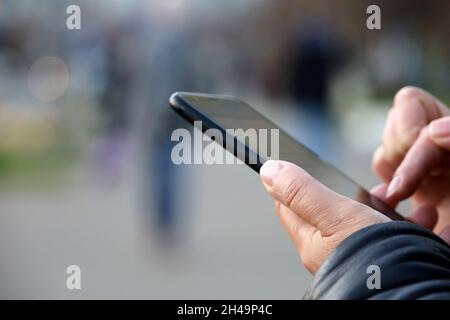 Mani femminili con smartphone in primo piano su sfondo sfocato della città. Donna che usa il telefono cellulare all'aperto, il concetto di sms, la comunicazione online Foto Stock