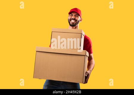 Servizio di consegna. Funny Middle Eastern Courier Man Holding Heavy pacchi scatole guardando la macchina fotografica in posa su sfondo giallo studio, indossando Foto Stock