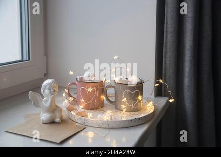 Decorazione appartamento per Natale. Tazze grigie e rosa con cuori bianchi dipinti con cacao, marshmallow su vassoio alla finestra, composizione decorati Foto Stock