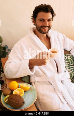 L'uomo ispanico beve il tè benessere durante il relax presso l'esotico centro benessere, indossando un accappatoio bianco e seduto su una poltrona Foto Stock