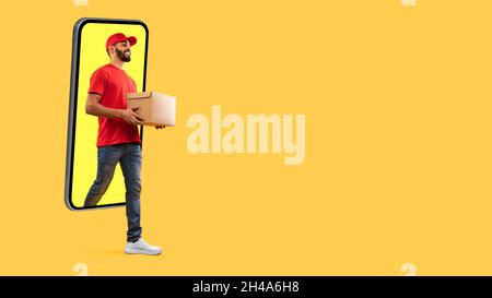 Middle Eastern Courier Man che esce dallo schermo dello smartphone trasportando la scatola che consegna il pacco sopra sfondo giallo dello studio. Concetto di applicazione di consegna. Foto Stock