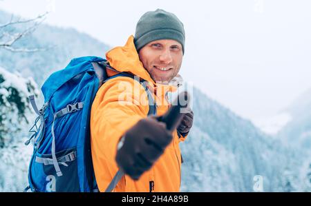 Uomo sorridente vestito giacca softshell arancione brillante con zaino guardando la fotocamera e mostrando un gesto di pollice-up mentre lui trekking montagna inverno Foto Stock