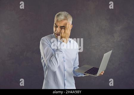 Uomo d'affari senior spaventato o timido che copre gli occhi guardando il laptop nelle mani. Foto Stock