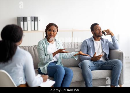 Trattamento di dipendenza del telefono cellulare. Infastidita donna afroamericana che lamenta l'uso eccessivo dello smartphone del marito durante la consultazione psicologica Foto Stock