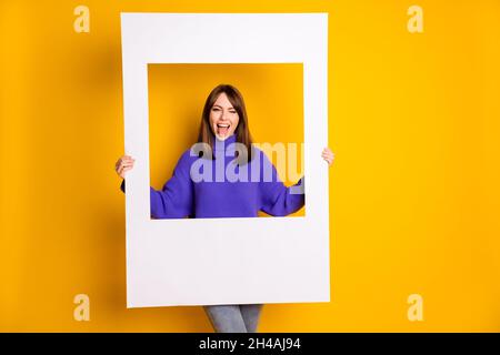 Foto di bella ragazza divertente vestito viola maglione tenendo carta foto cornice verricello lingua isolato giallo sfondo di colore Foto Stock