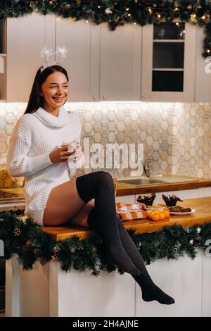 Una ragazza a Natale è seduta sul tavolo in cucina, tenendo una torta e sognando.Una donna alla vigilia di Capodanno in cucina. Foto Stock