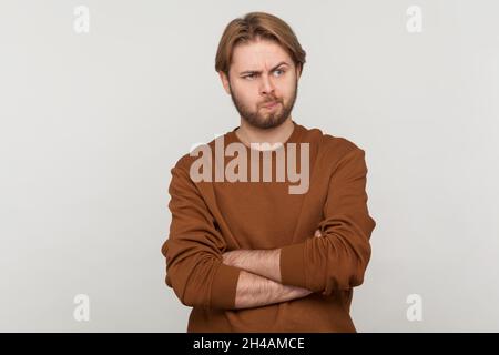 Ritratto di bell'uomo con barba che indossa una felpa, in piedi, braccia incrociate, guardando via e pensando a cosa fare, confuso non sicuro di soluzione. Studio interno girato isolato su sfondo grigio. Foto Stock