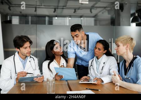 Gruppo multietnico concentrato di medici uomini e donne in abbigliamento da lavoro seduti a tavola in clinica, avendo breefing, cercando soluzioni, discutendo c Foto Stock