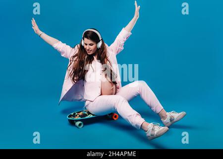 una donna incinta in vestito rosa si siede su uno skateboard con cuffie e ascolta musica su sfondo blu. Foto Stock