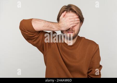 Ritratto di uomo con barba indossare la felpa, chiudere gli occhi con la mano, non vuole vedere che, ignorando i problemi, nascondendosi da situazioni stressanti. Studio interno girato isolato su sfondo grigio. Foto Stock