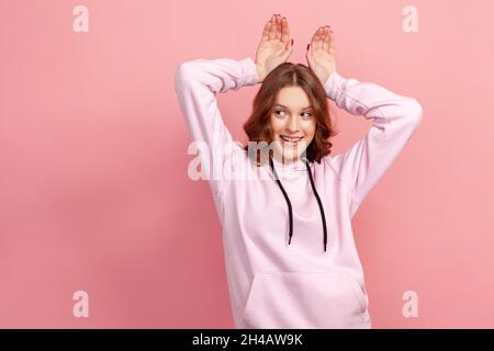 Ritratto di spensierato felice brunette giovane femmina in felpa con cappuccio facendo divertente coniglietto orecchie con le mani sulla testa, comportamento infantile, allegro umore ottimista. Studio interno girato isolato su sfondo rosa Foto Stock