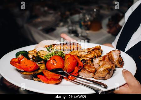 La sera, il ristorante serve un piatto caldo di barbecue con vegetables.fried carni con pomodori e altre verdure su un piatto in un ristorante Foto Stock