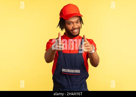 Ritratto di positivo bearded costruttore o corriere indossando tute, puntando a macchina fotografica con le dita, guardando con piacevole sorriso, servizio. Studio interno girato isolato su sfondo giallo. Foto Stock