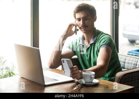 Ritratto di un bel uomo d'affari occupato indossando una T-shirt verde, lavorando su un laptop, parlando sul cellulare e guardando un altro cellulare. Foto al coperto vicino alla grande finestra, sfondo del caffè. Foto Stock