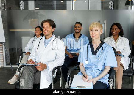 Il gruppo multirazziale di medici ha concentrato uomini e donne in abbigliamento da lavoro frequentando una conferenza medica in clinica, seduto su sedie e guardando copia spac Foto Stock