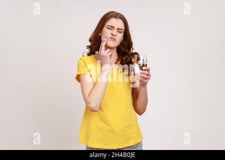 Ritratto di giovane donna malata triste con capelli ricci in piedi e toccare la sua guancia perché sensazione di dolore sul dente, dopo aver bevuto acqua fredda. Studio interno girato isolato su sfondo grigio. Foto Stock