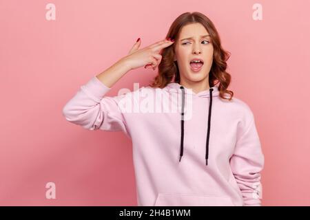 Ritratto di depressa ricci capelli ragazza teen in hoodie puntando dito pistola alla testa e cercando disperato, facendo suicidio gesto, sparando se stessa. Studio interno girato isolato su sfondo rosa Foto Stock