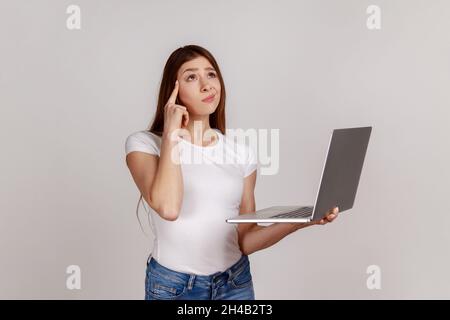 Ritratto di confuso puzzled donna pensierosa che tiene il laptop pensando alla domanda difficile, soluzione ponderante, indossando la T-shirt bianca. Studio interno girato isolato su sfondo grigio. Foto Stock