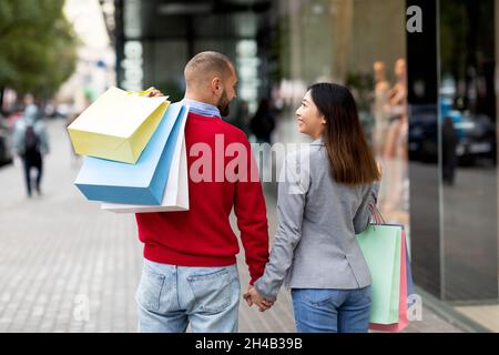 Vista posteriore della giovane coppia internazionale che cammina mano a mano vicino al grande centro commerciale, tenendo i sacchetti per gli acquisti all'aperto. Famiglia multirazziale millenario shoppi finestra Foto Stock