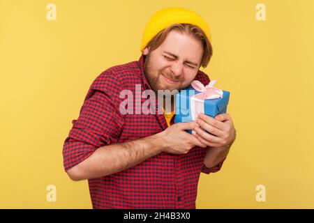 Ritratto di hipster felice ported ragazzo in berretto e camicia a scacchi in piedi e tenendo presente scatola e abbracciare, regalo da tempo atteso. Studio interno girato isolato su sfondo giallo. Foto Stock