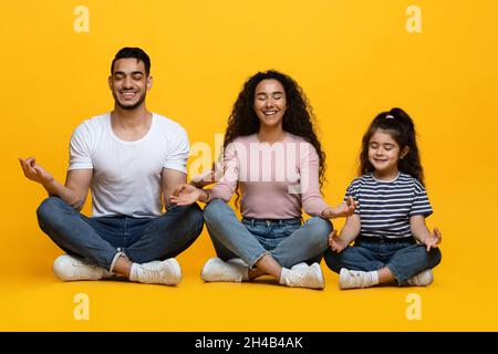 Happy Arab Famiglia di tre con bambina meditating insieme in Studio, sorridendo genitori del Medio Oriente e bambino seduto in Lotus posizione sopra Foto Stock