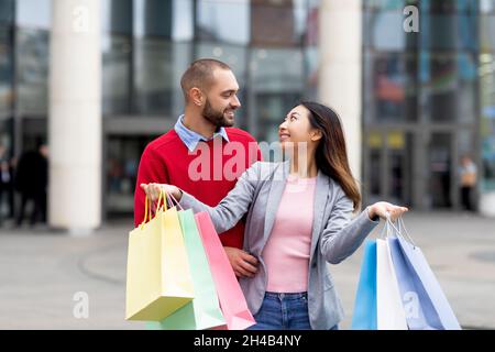Affettuoso giovane coppia interrazziale in piedi per strada con borse shopping, guardando l'un l'altro, soddisfatto dei loro acquisti, in posa vicino alla città m Foto Stock