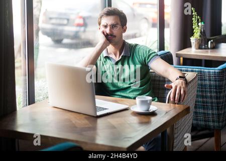 Ritratto di un dipendente uomo che indossa una T-shirt verde, guarda la fotocamera con espressione seria, tenere la mano sotto il mento, lavorare online su un notebook, scatto interno vicino a una grande finestra, sfondo caffè. Foto Stock