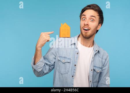 Ritratto di stupito uomo indossando una camicia in denim, in piedi puntando verso la casa di carta sulla spalla e sorridendo alla macchina fotografica, acquisto a casa. Studio interno girato isolato su sfondo blu. Foto Stock