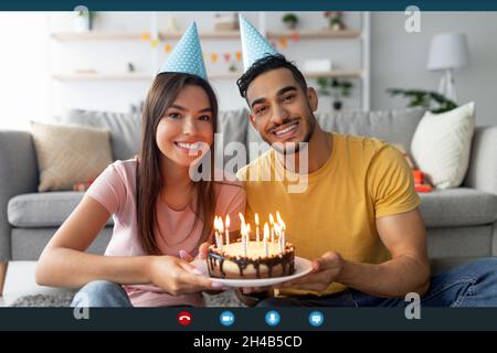 Schermo del computer di felice coppia multirazziale in cappelli di partito mostrando torta di compleanno con candele alla webcam, celebrando il b-day online, facendo video ca Foto Stock