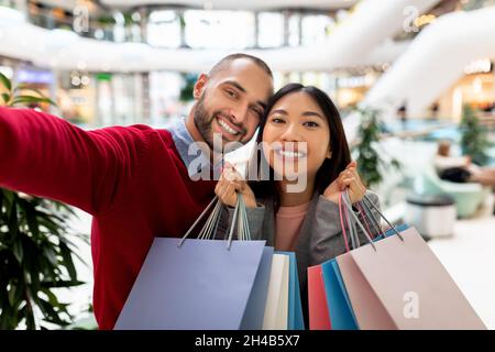 Bella coppia allegra interrazziale che tiene sacchetti di carta, prendendo selfie al centro commerciale, andando a fare shopping insieme. Amorevole coniugi multinazionali che acquistano bene Foto Stock