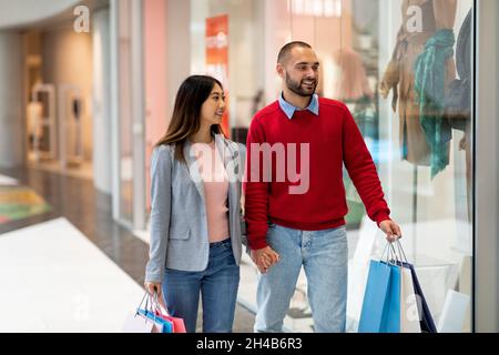 Amorevole coppia interracial camminare lungo le finestre del deposito, le mani di tenuta, shopping per i vestiti al centro commerciale, spazio di copia. Coniugi millenni diversi con carta Foto Stock