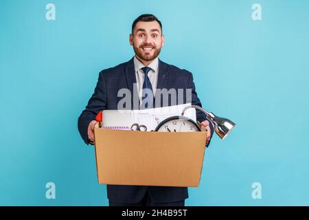 Estremamente felice giovane uomo adulto con barba che indossa abito scuro stile ufficiale, si alza sorridente tenendo grande scatola di cartone con il suo roba, ottenendo nuovo lavoro. Studio interno girato isolato su sfondo blu. Foto Stock
