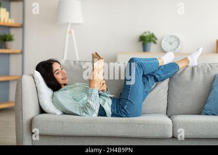 Passatempi di soggiorno a casa. Donna indiana millenaria sdraiata sul divano con libro aperto al coperto, godendo tranquilla mattina del fine settimana. Giovane donna orientale che legge capti Foto Stock