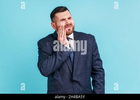 Uomo infelice indossato vestito ufficiale stile sensazione di mal di denti, toccare guancia dolorosa, soffrire di cavità, denti incrinati, recessione gengivale. Studio interno girato isolato su sfondo blu. Foto Stock