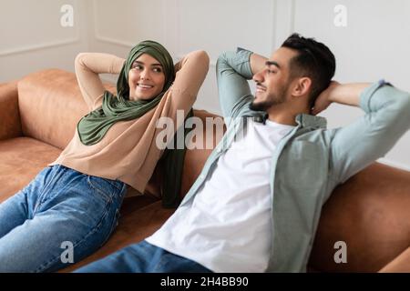 Felice Medio Oriente coppia appoggiarsi indietro sul divano mentre si rilassa a casa insieme, giovane ragazzo arabo e la signora in Headscarf riposo in accogliente soggiorno, S Foto Stock