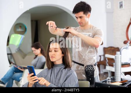 Ragazza cliente utilizzando il telefono durante il taglio dei capelli da professionale parrucchiere Foto Stock